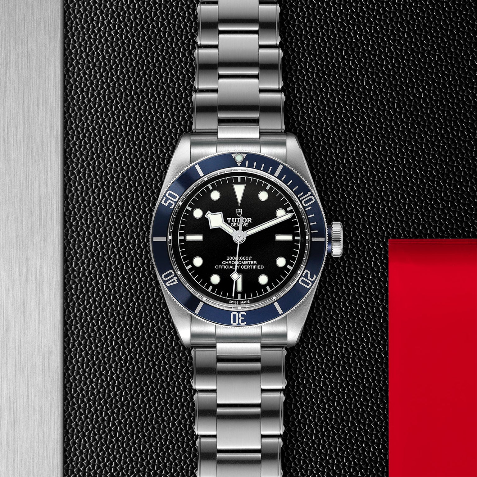A TUDOR Black Bay wristwatch with a stainless steel bracelet and black dial is set against a textured black background with silver and red accents.