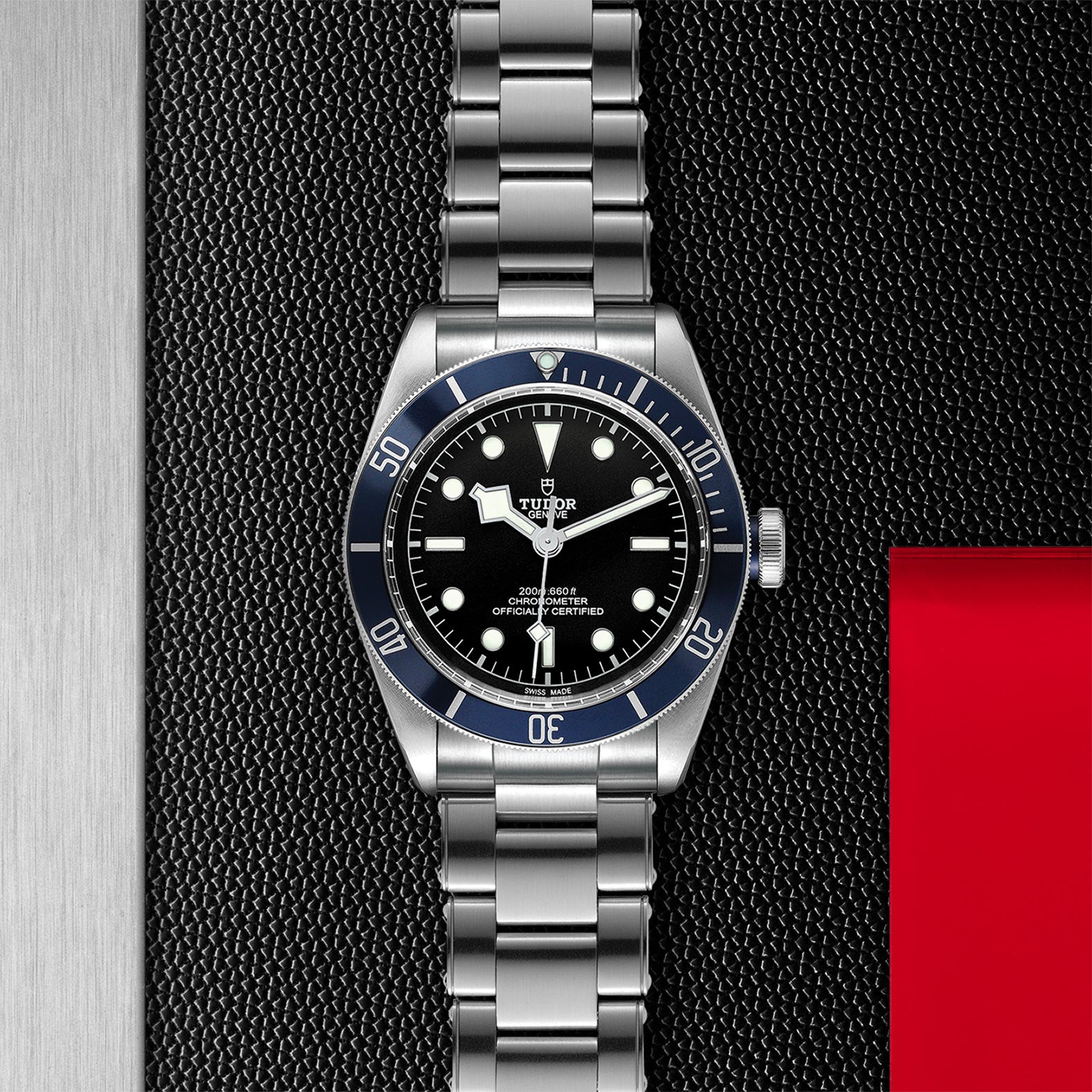 A TUDOR Black Bay wristwatch with a stainless steel bracelet and black dial is set against a textured black background with silver and red accents.