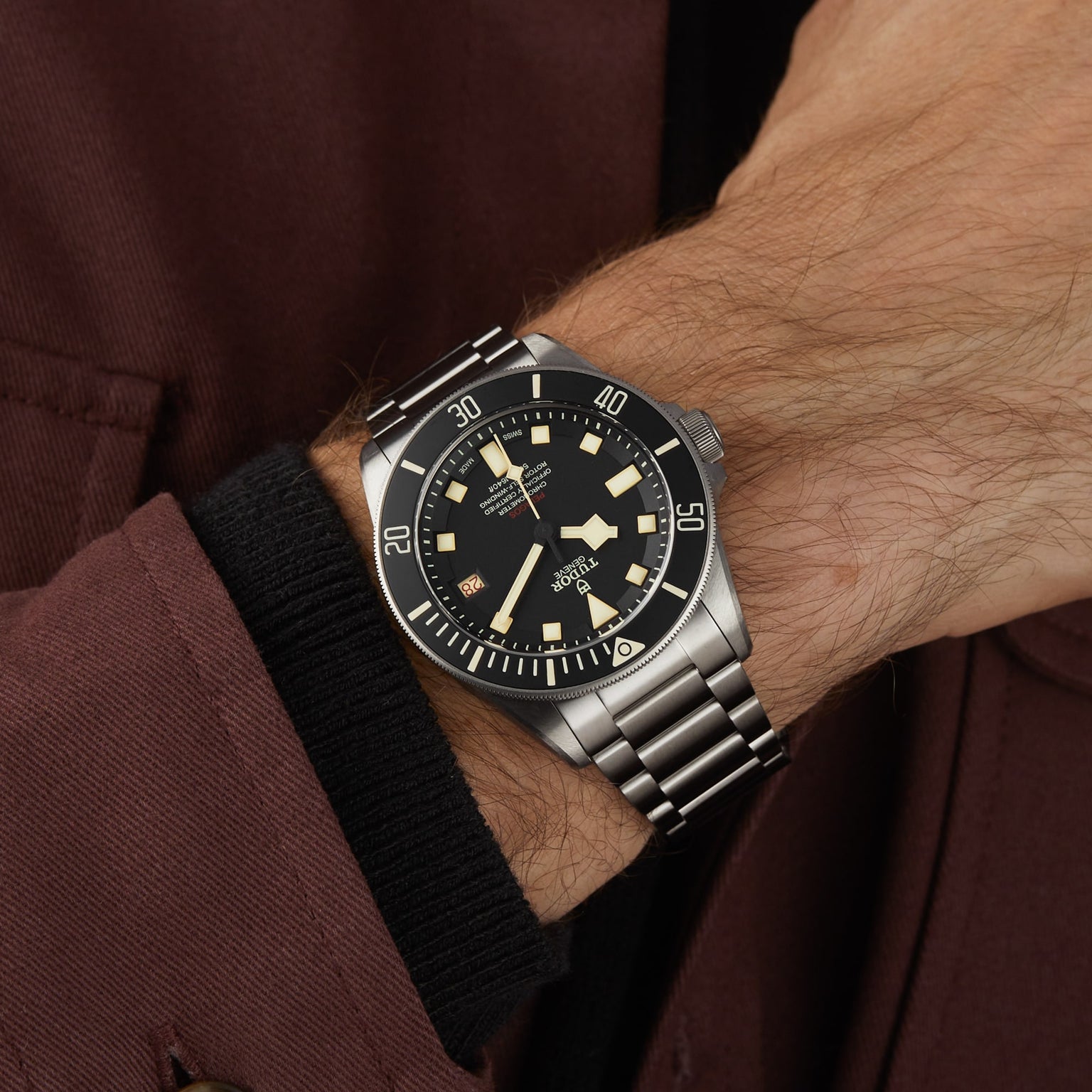 The TUDOR Pelagos LHD, a stainless steel diver’s watch with a black bezel and dial, is worn on the left wrist and partially concealed by a maroon coat sleeve.