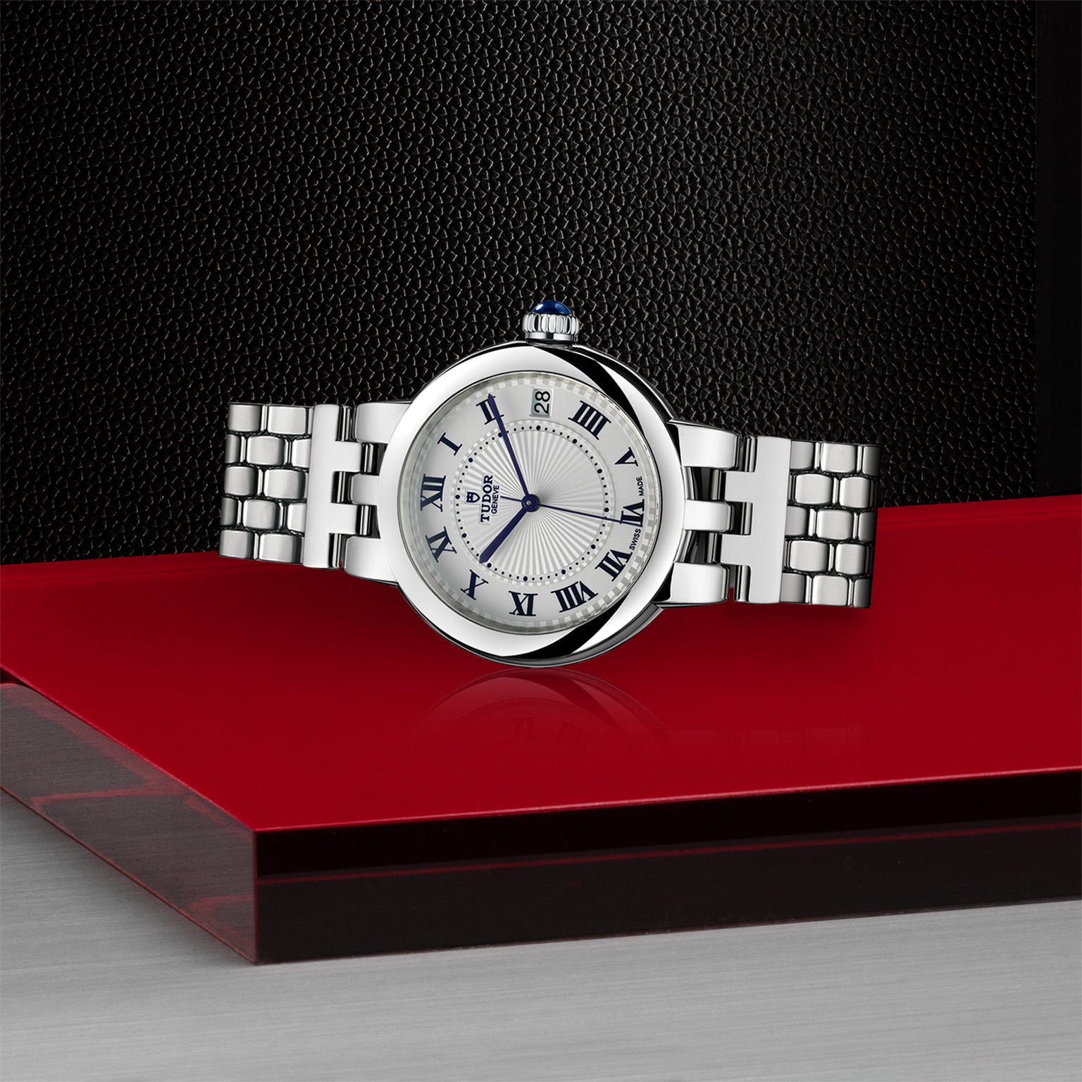 The TUDOR Clair de Rose silver wristwatch with Roman numerals rests on a red and gray surface, set against a black textured background.