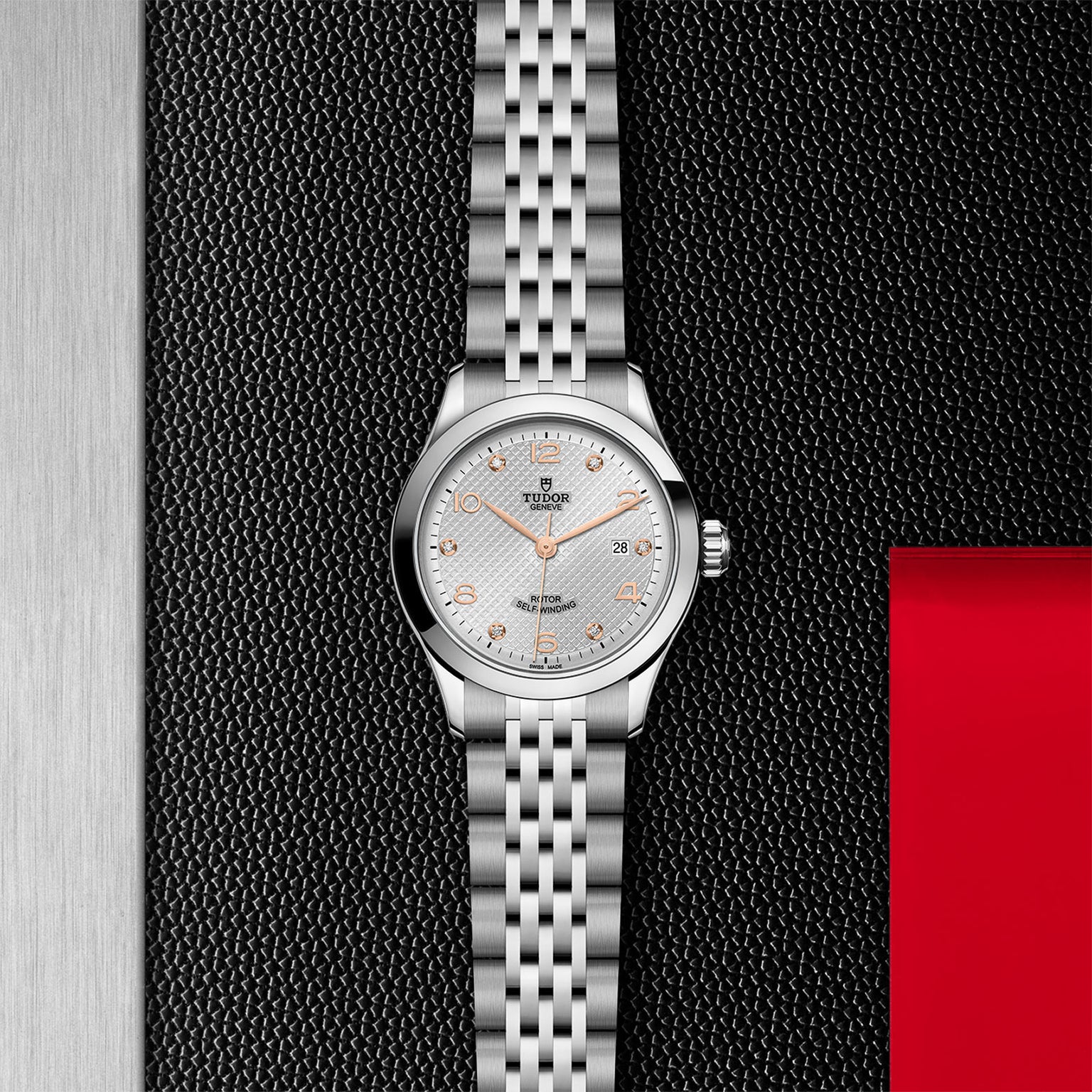 A silver TUDOR 1926 wristwatch with a metal link band and white textured dial is shown upright against a black and red textured background.