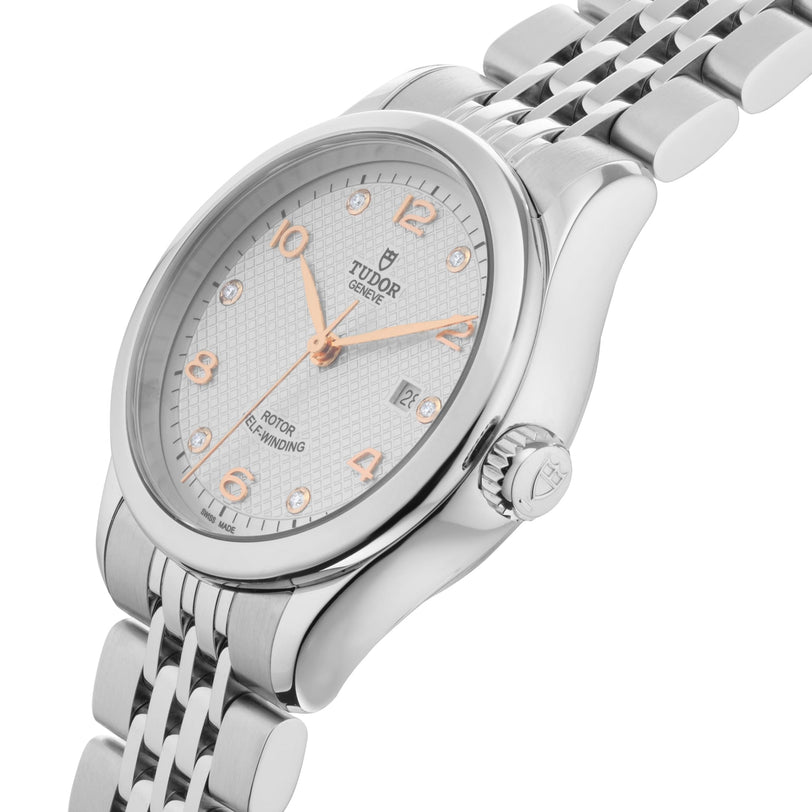 The TUDOR 1926 is a silver wristwatch with a metal link band, white textured dial, rose gold numerals, diamond hour markers, date display, and refined self-winding movement, shown on a white background.