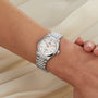 A close-up of a person’s wrist wearing a silver TUDOR 1926 watch with a white dial and gold numerals, featuring self-winding movement, set against a cream fabric background.