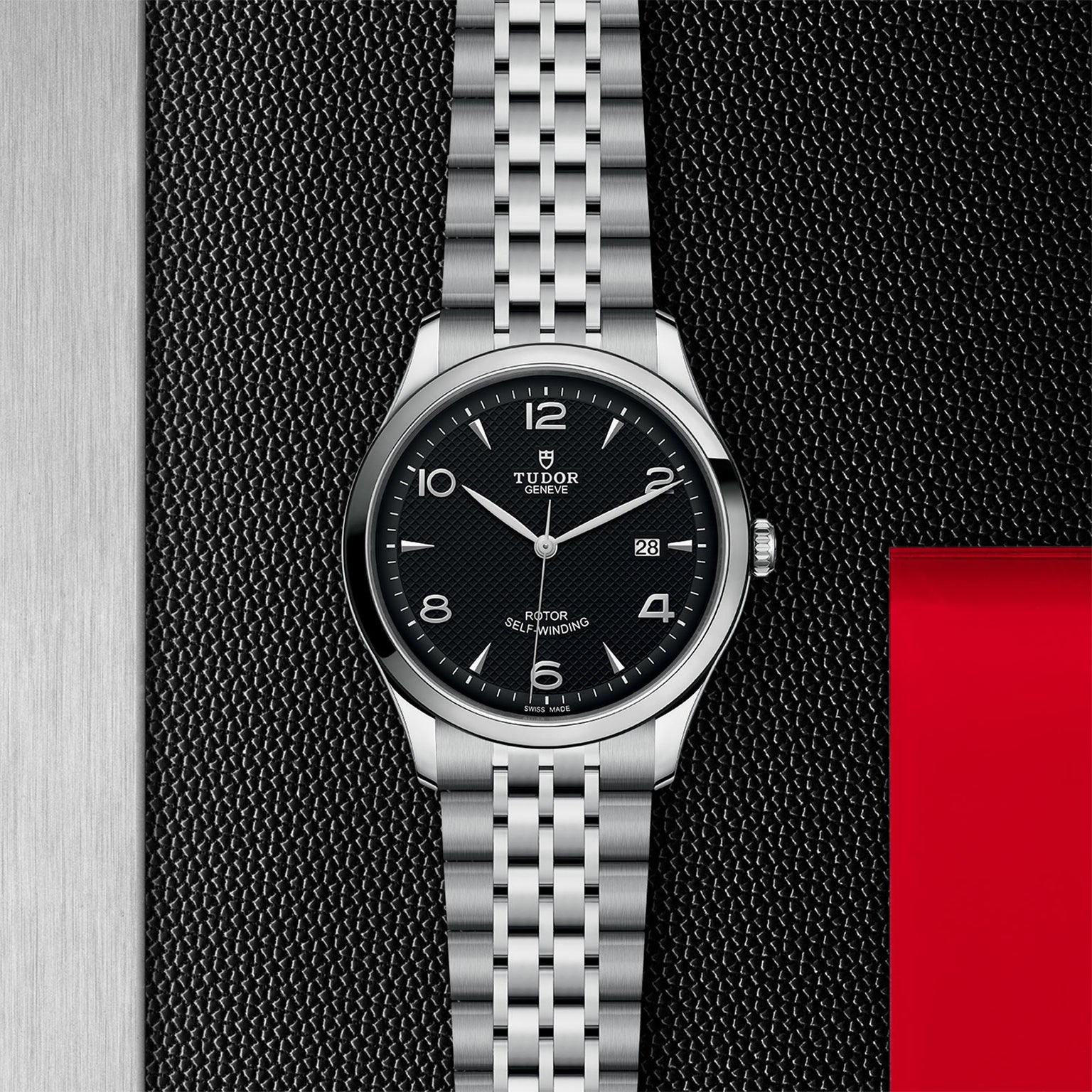 The TUDOR 1926 wristwatch features a black dial, silver hour markers, date display at 3 o’clock, and a silver metal bracelet with sapphire crystal, shown against a textured black and red background.