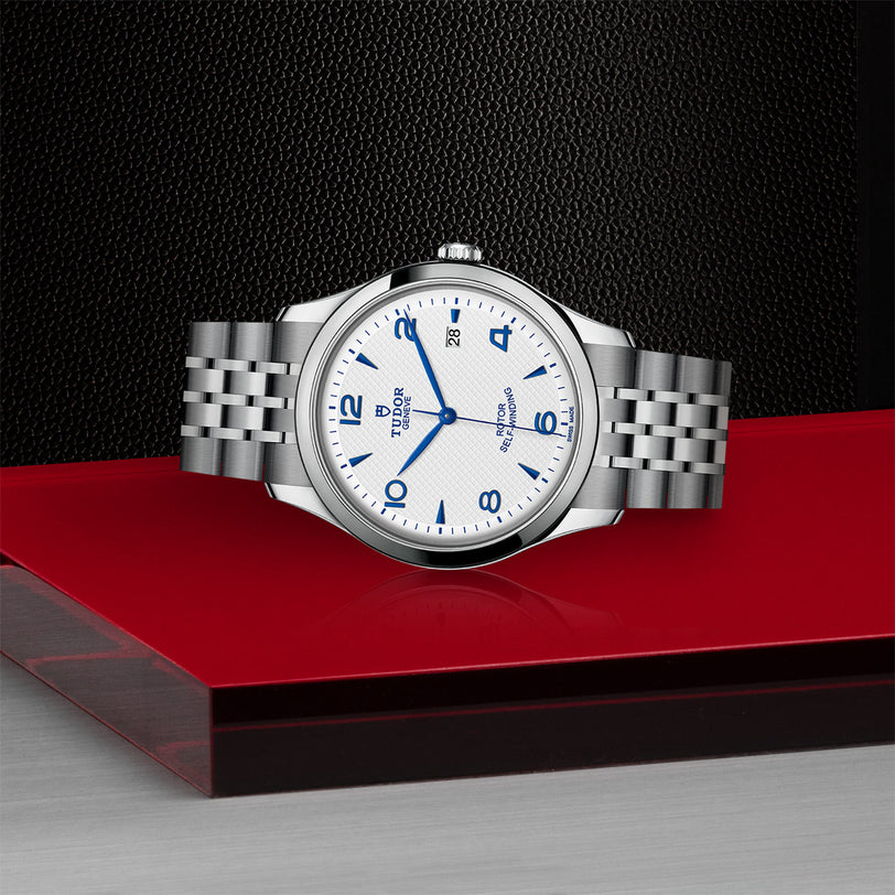 A TUDOR 1926 mens watch with a stainless steel bracelet, white dial, and blue numerals is showcased on a red and black surface against a textured black background—perfect for those who appreciate classic mechanical timepieces.