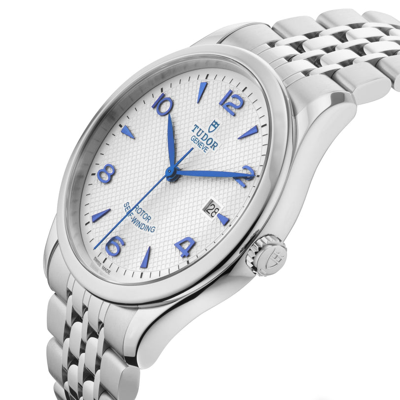 The TUDOR 1926 wristwatch features a silver case, white textured dial, blue hands, date window, and metal bracelet.