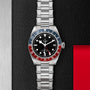 A TUDOR Black Bay GMT wristwatch with a stainless steel bracelet, black dial, and red-blue bezel is shown on a textured black and metallic surface.