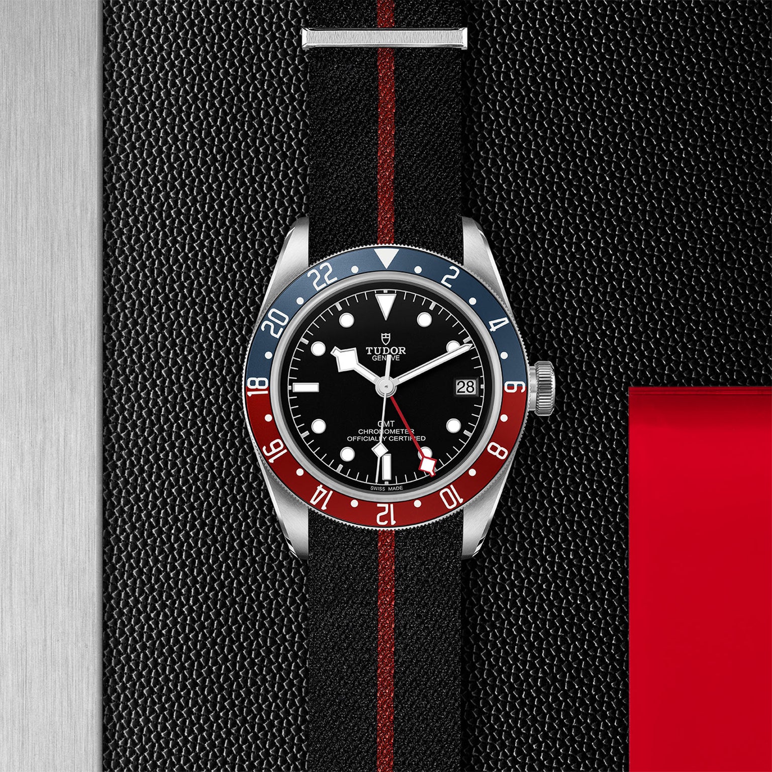 A TUDOR Black Bay GMT wristwatch with a black dial, blue and red bezel, and a black fabric strap with red stripe rests on a textured black surface with metallic and red accents, reflecting classic diving watch style.