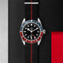 A TUDOR Black Bay GMT wristwatch with a black dial, blue and red bezel, and a black fabric strap with red stripe rests on a textured black surface with metallic and red accents, reflecting classic diving watch style.