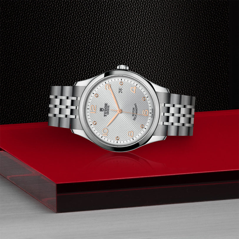 A TUDOR 1926 silver wristwatch with a round white dial, gold hour markers, and metal link band is displayed on a red and black surface against a textured black background.