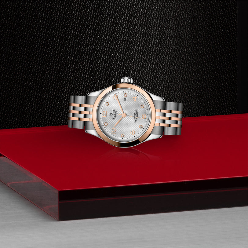 The TUDOR 1926 watch, featuring a rose gold bezel and patterned dial, is displayed on a glossy red and gray surface with a textured black background.