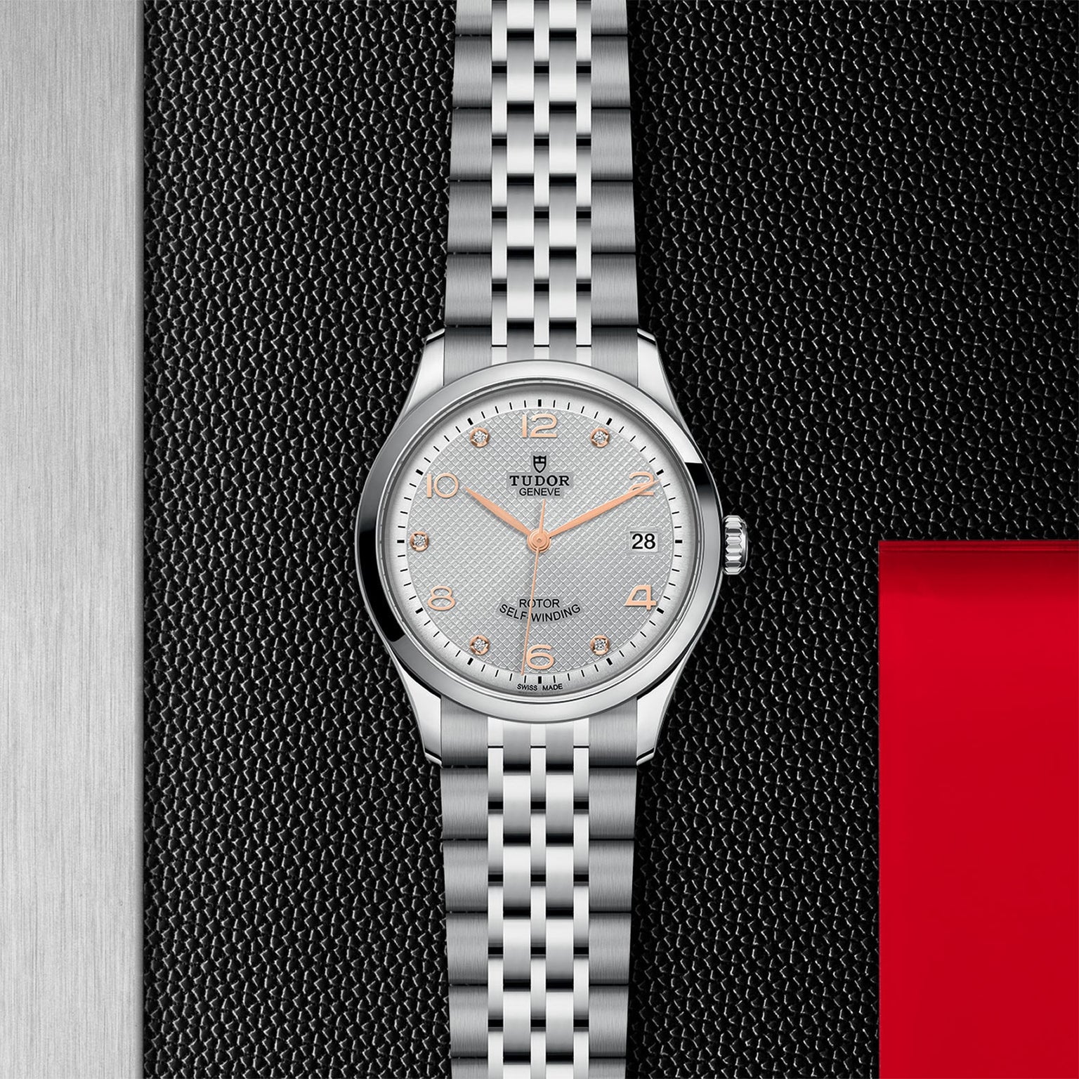 A silver TUDOR 1926 wristwatch with a metal bracelet, white textured dial, gold hour markers, and date display rests on a black surface with red and metallic accents—a classic mechanical timepiece.