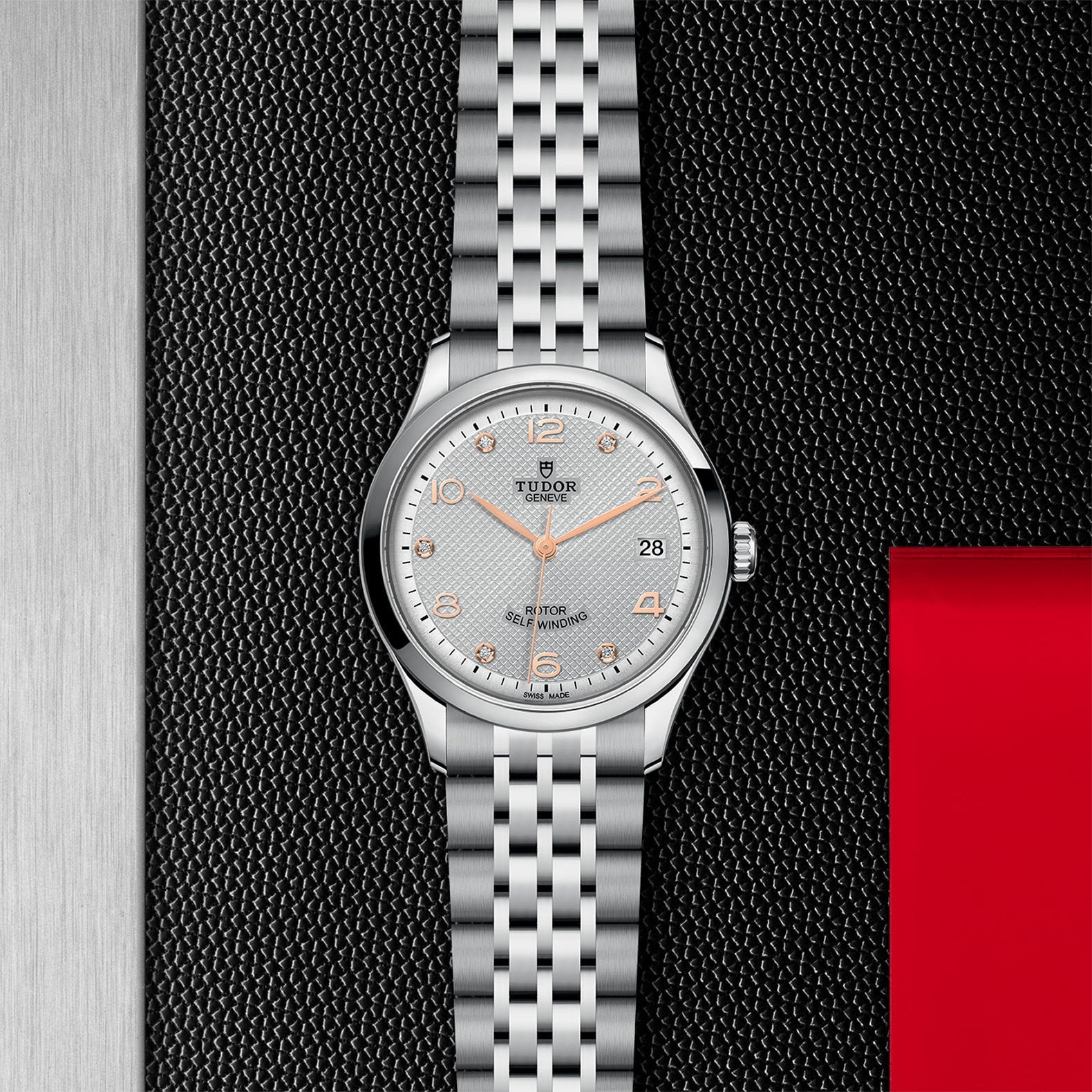 A silver TUDOR 1926 wristwatch with a metal bracelet, white textured dial, gold hour markers, and date display rests on a black surface with red and metallic accents—a classic mechanical timepiece.