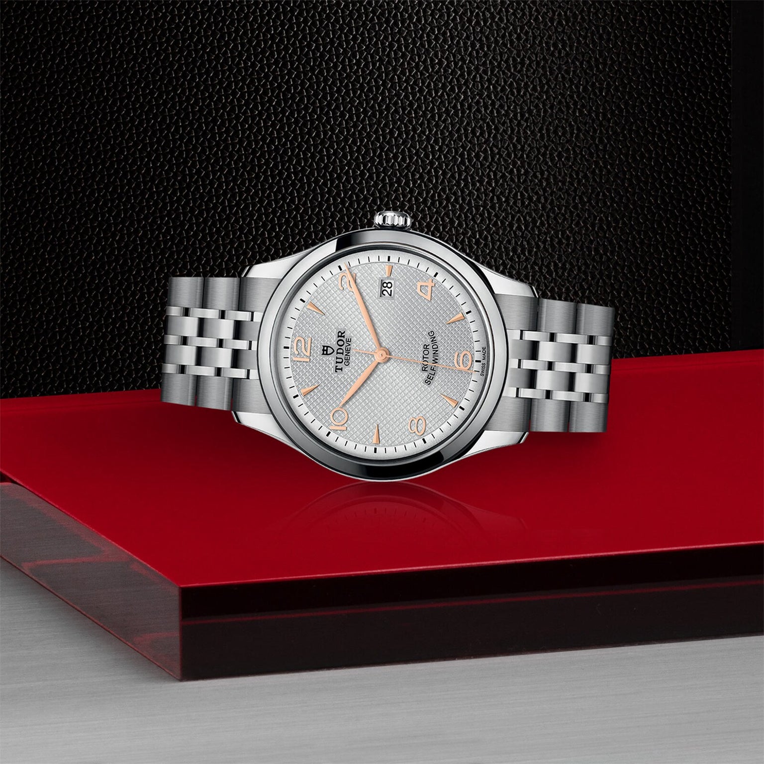 A classic and elegant TUDOR 1926 silver wristwatch with a white textured dial and metal bracelet is displayed on a red and black platform against a textured black background.