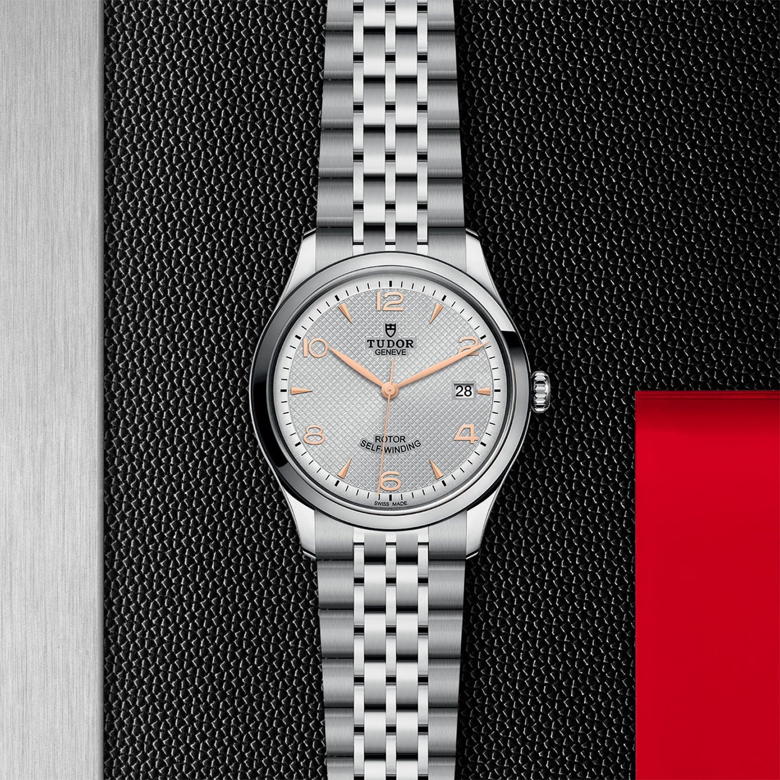 A classic and elegant TUDOR 1926 watch in stainless steel with a silver dial, orange hour markers, date at 3 oclock, and a five-link bracelet, shown on a textured black surface with a red accent.