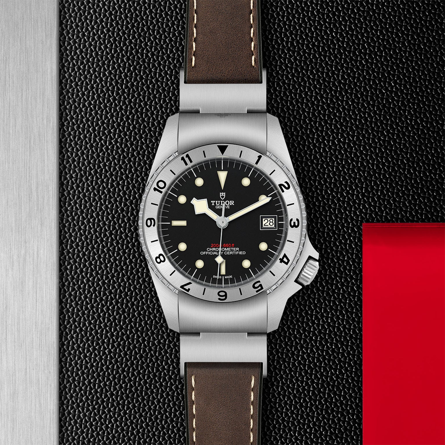 The TUDOR Black Bay P01 wristwatch, with signature snowflake hands, a brown leather strap, black dial, silver bezel, and date display, is displayed on a textured black and red surface.