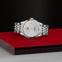 A silver TUDOR 1926 wristwatch with a metal bracelet and textured white dial rests on a glossy red and gray surface against a black backdrop, highlighting the classic elegance of TUDOR’s mechanical craftsmanship.