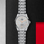A TUDOR 1926 silver watch with orange-accented face and metal band is displayed on a textured black surface.