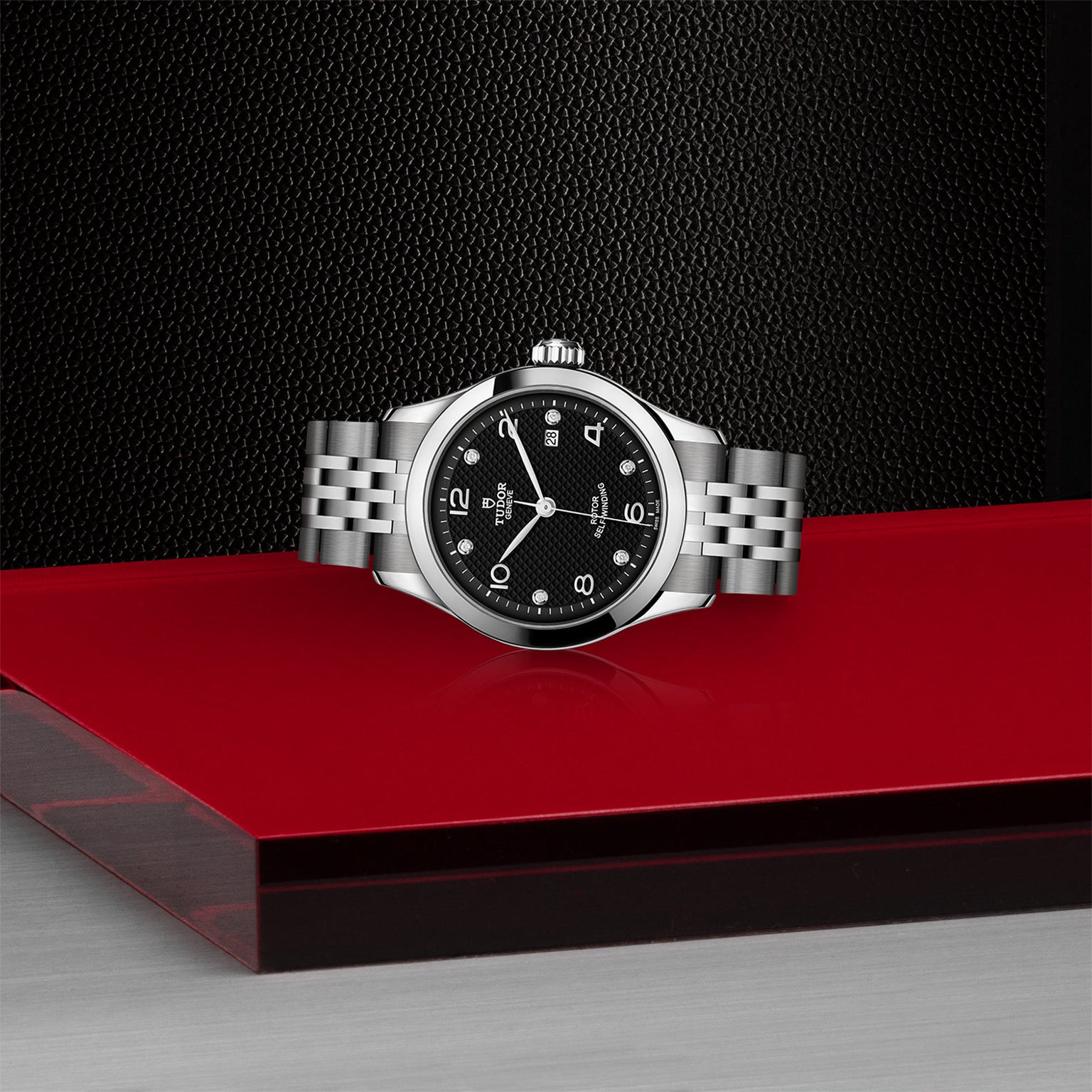 The TUDOR 1926, a timelessly elegant silver wristwatch with a black face and metal band, is showcased on a red and silver platform against a textured black background.