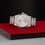 A TUDOR 1926 silver and rose gold wristwatch with a textured white dial is showcased on a red and black platform against a dark textured background.