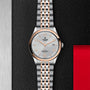 A TUDOR 1926 wristwatch with a silver and rose gold bracelet is displayed on a textured black surface.