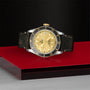 The TUDOR Black Bay S&G wristwatch features a gold dial, black bezel, and distressed black leather strap, elegantly displayed on a glossy red platform set against a textured black background.