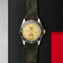 The TUDOR Black Bay S&G features a gold dial, black bezel, and distressed black leather strap—an iconic timepiece displayed on a textured black and red surface.