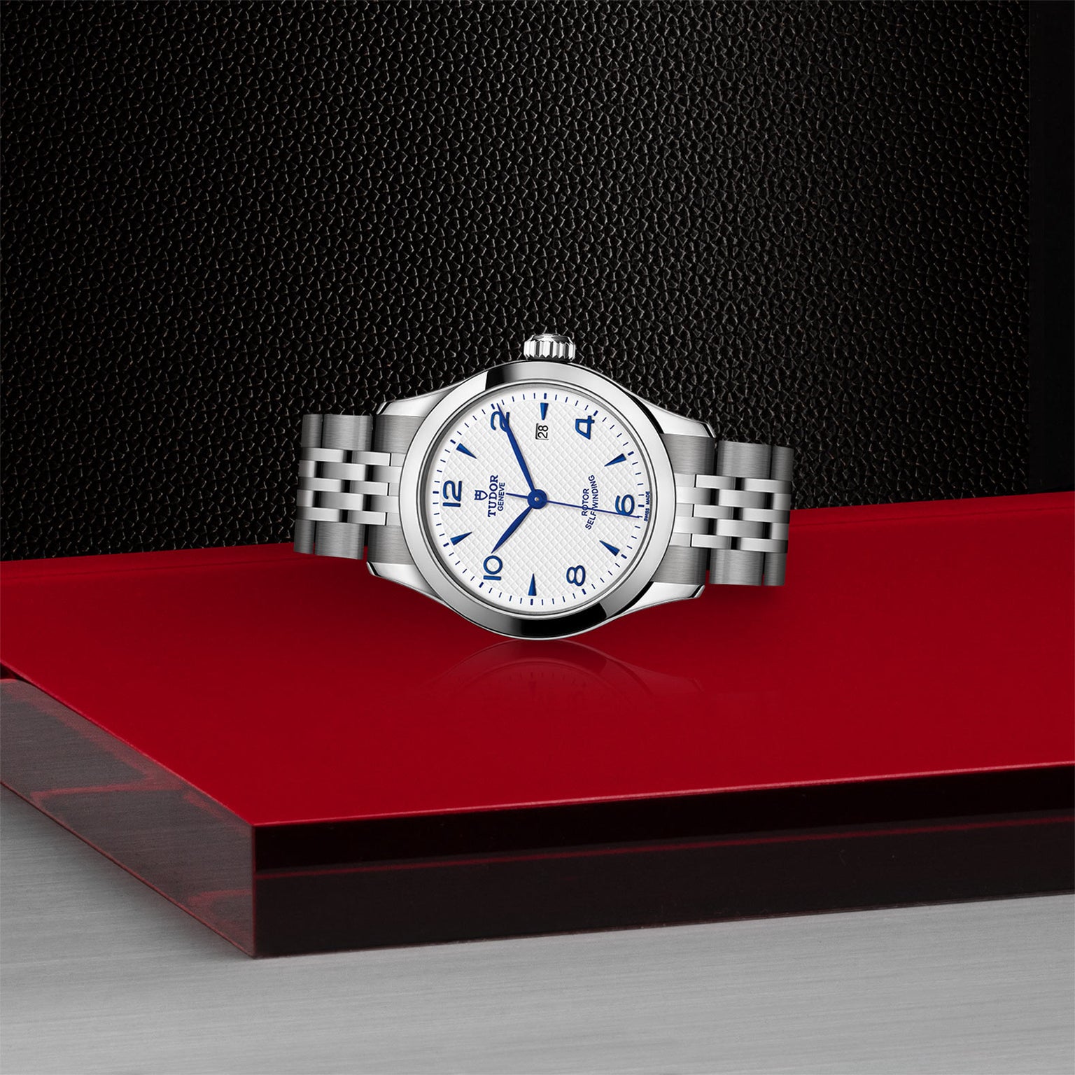 The TUDOR 1926 silver wristwatch features a white face with blue numerals, shown on a glossy red and gray surface.