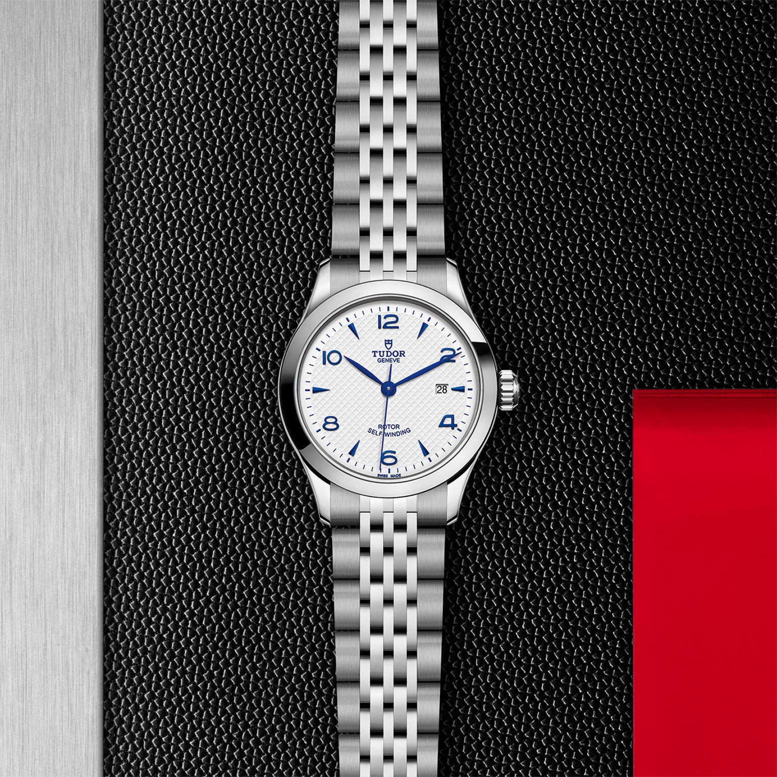 A silver TUDOR 1926 wristwatch with a white textured dial, blue numerals, date window, and metal bracelet is displayed on a black and red background—a refined pick for mechanical watch enthusiasts.