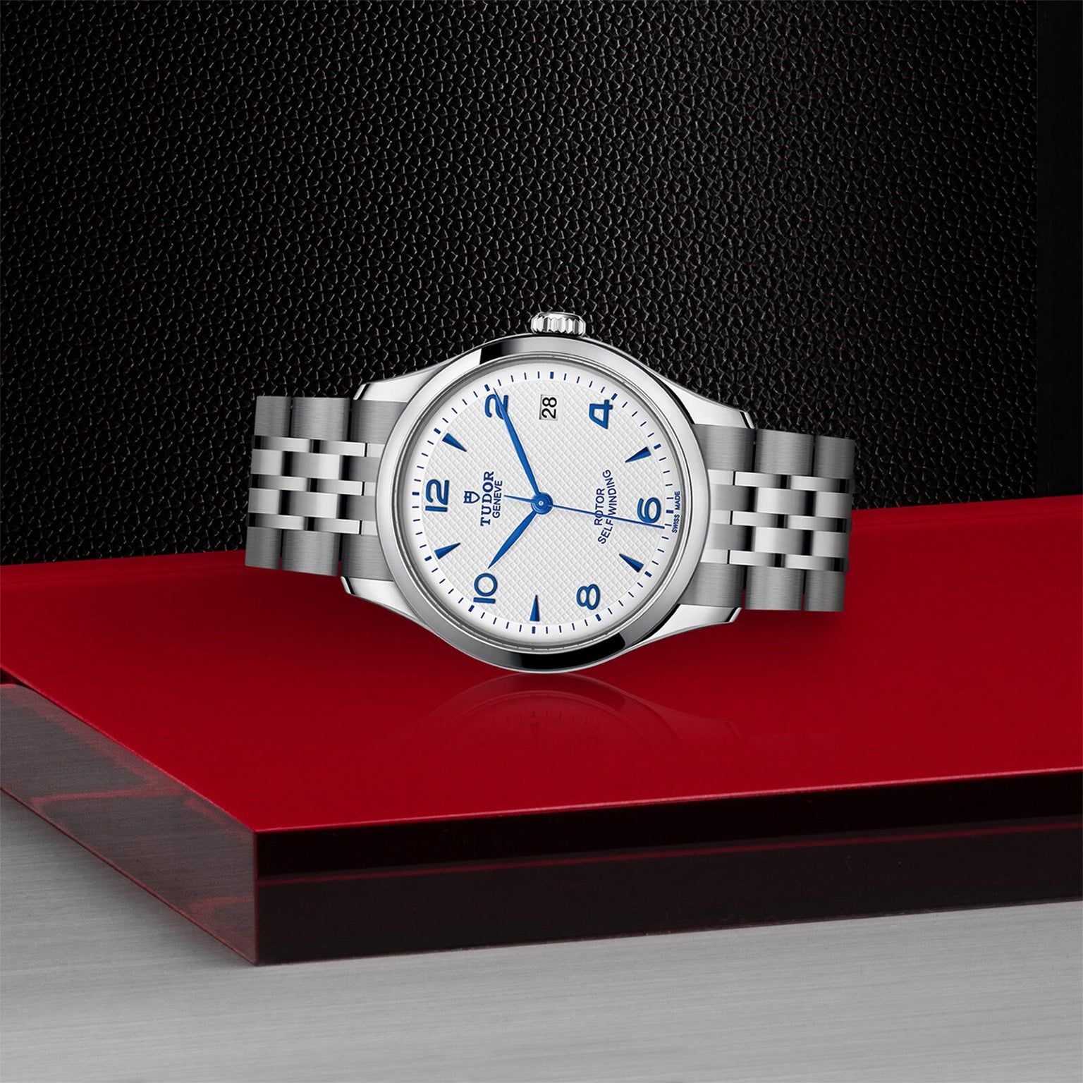 A silver TUDOR 1926 wristwatch with a white dial, blue numerals, and date display rests on a red surface with a black textured and silver background.