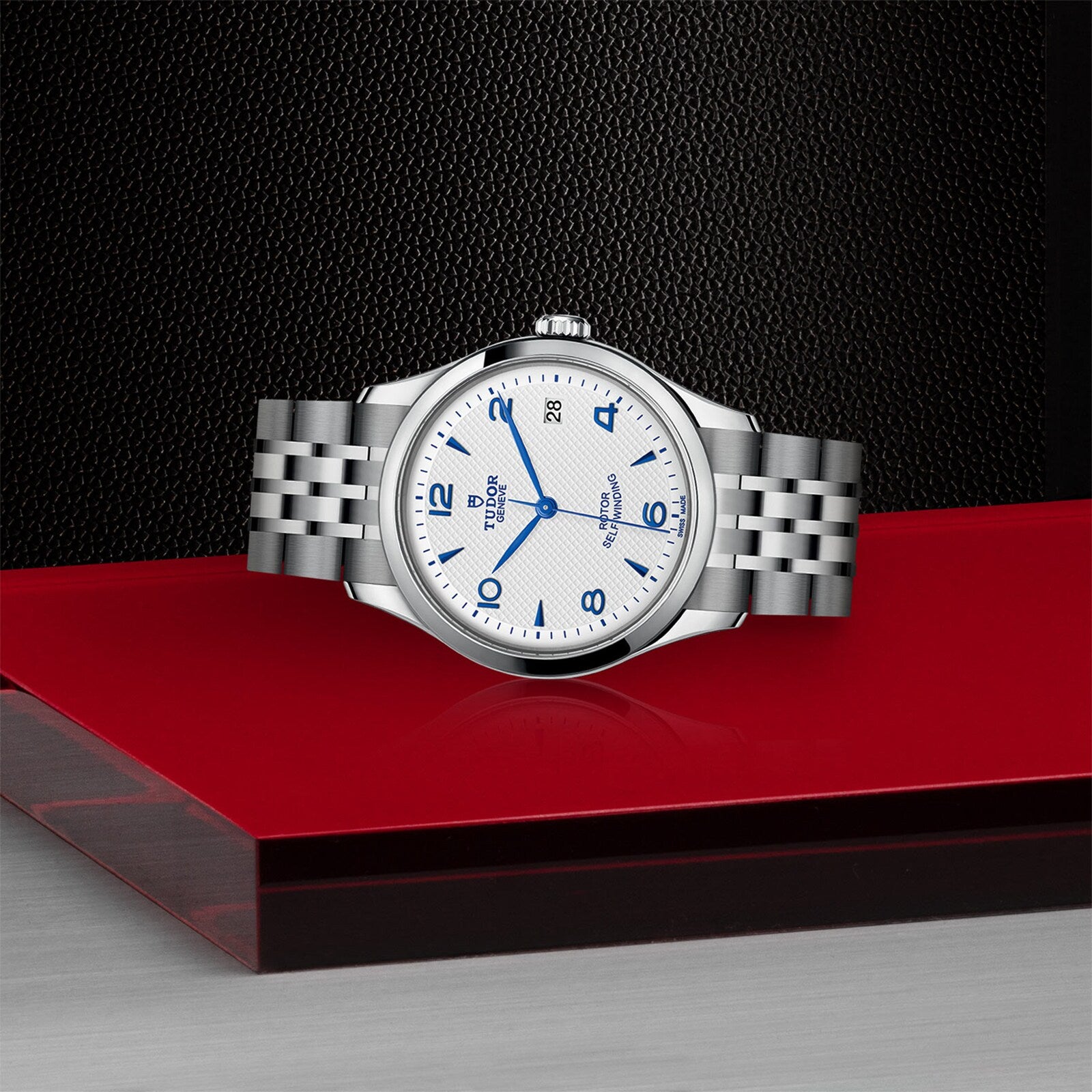 A silver TUDOR 1926 wristwatch with a white dial, blue numerals, and date display rests on a red surface with a black textured and silver background.