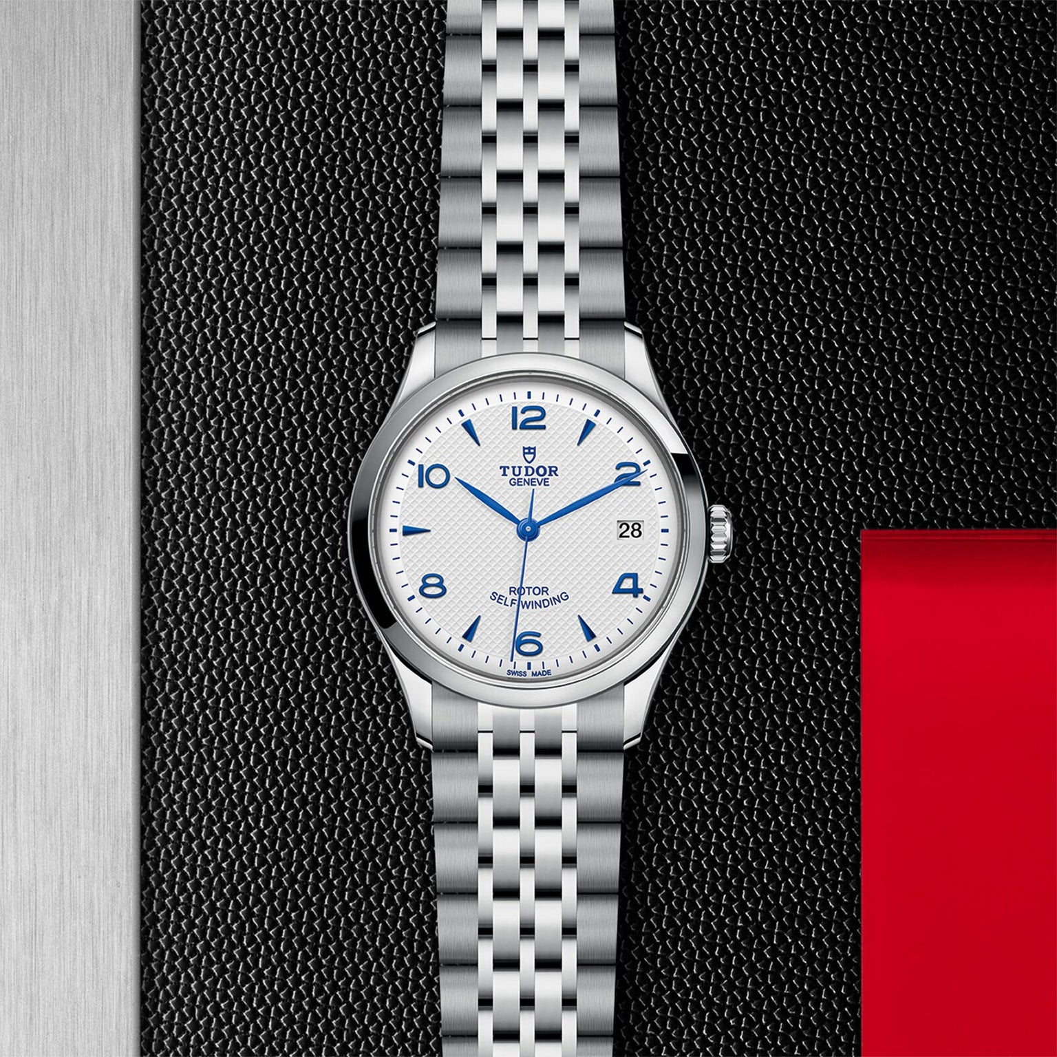 The TUDOR 1926 wristwatch, featuring a silver metal bracelet, white dial with blue numerals, and date display, rests on a textured black and red surface—a timeless classic for discerning watch enthusiasts.