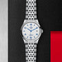 The TUDOR 1926 wristwatch, featuring a silver metal bracelet, white dial with blue numerals, and date display, rests on a textured black and red surface—a timeless classic for discerning watch enthusiasts.