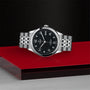 The TUDOR 1926 silver wristwatch features a black dial and metal band, displayed on a red and black surface.
