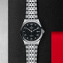 The TUDOR 1926 wristwatch features a black dial, silver hour markers, and a silver metal bracelet, showcased on a textured black and metallic background with a red accent—celebrating TUDORs heritage in mechanical watchmaking.