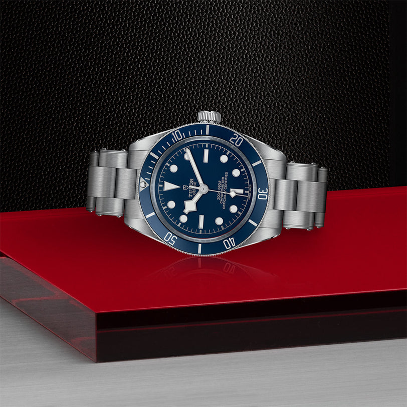 A TUDOR Black Bay 58 watch with a blue dial and bezel is showcased on a red and black geometric surface, emphasizing its iconic design inspired by classic diving watches.