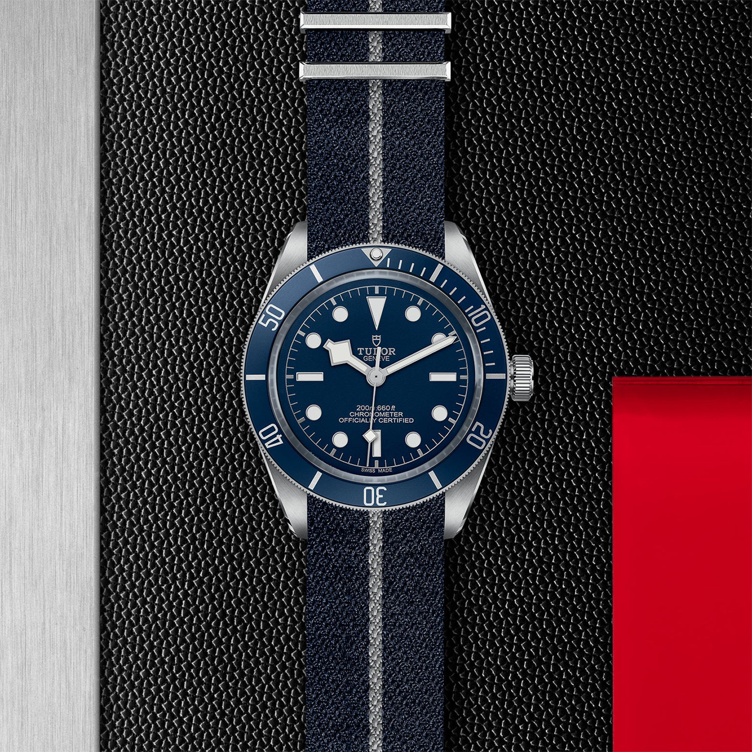 The TUDOR Black Bay 58, a blue diving watch on a striped fabric strap, is shown on a textured black surface with silver metal and red accents, highlighting its self-winding mechanical movement.