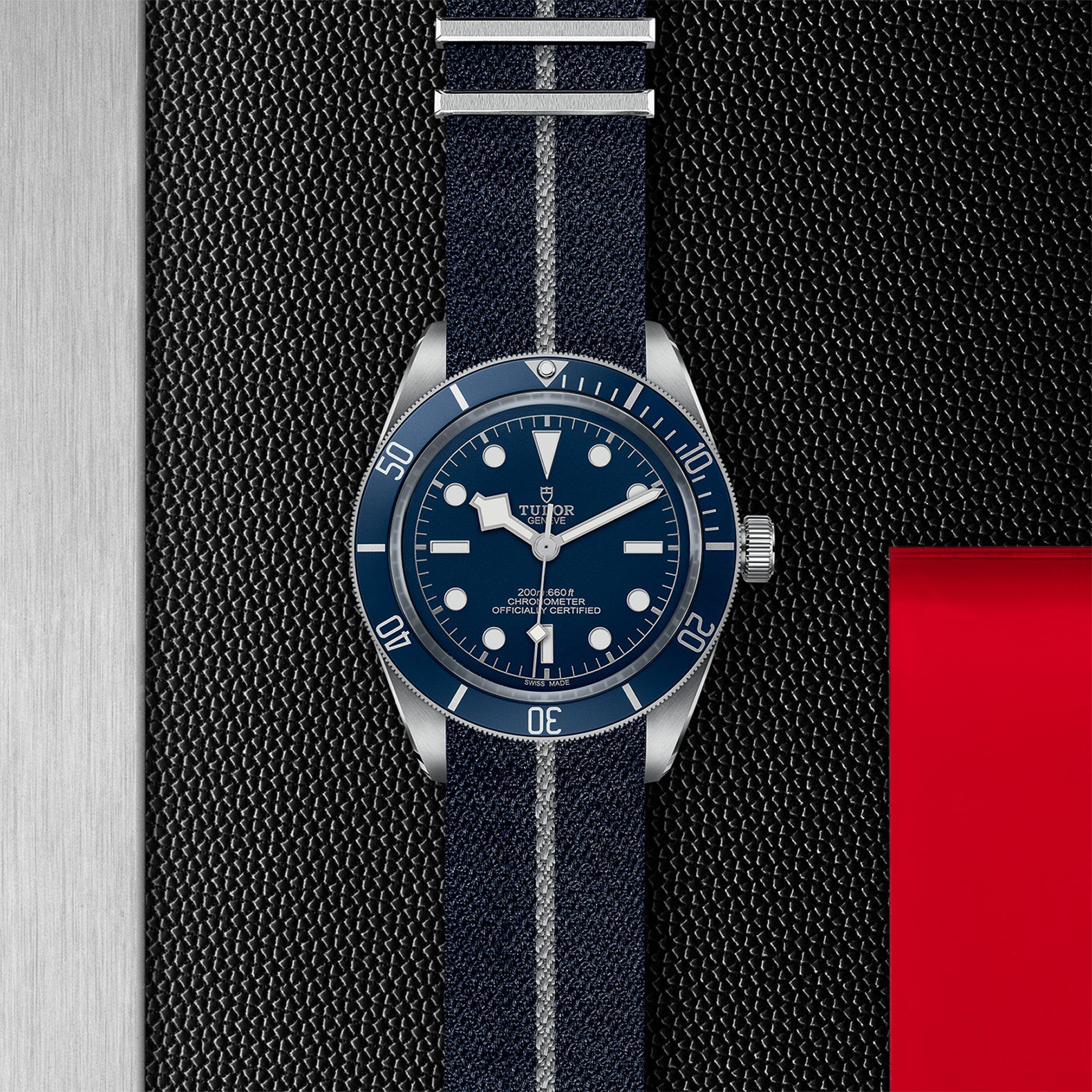 The TUDOR Black Bay 58, a blue diving watch on a striped fabric strap, is shown on a textured black surface with silver metal and red accents, highlighting its self-winding mechanical movement.