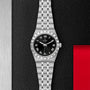 The TUDOR Royal wristwatch features a black dial, Roman numerals, date window, and a metal link bracelet. Displayed on a textured black background, it embodies the refined elegance of sport-chic design.