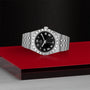 The TUDOR Royal wristwatch features a black dial, Roman numerals, and date window—an ideal choice for sport-chic enthusiasts. Shown on a striking red and black surface.