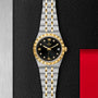 The TUDOR Royal features a black dial with gold Roman numerals and a date display at 3 o’clock, highlighted against a textured black and metallic background—embodying the sport-chic style and technical excellence of TUDOR.
