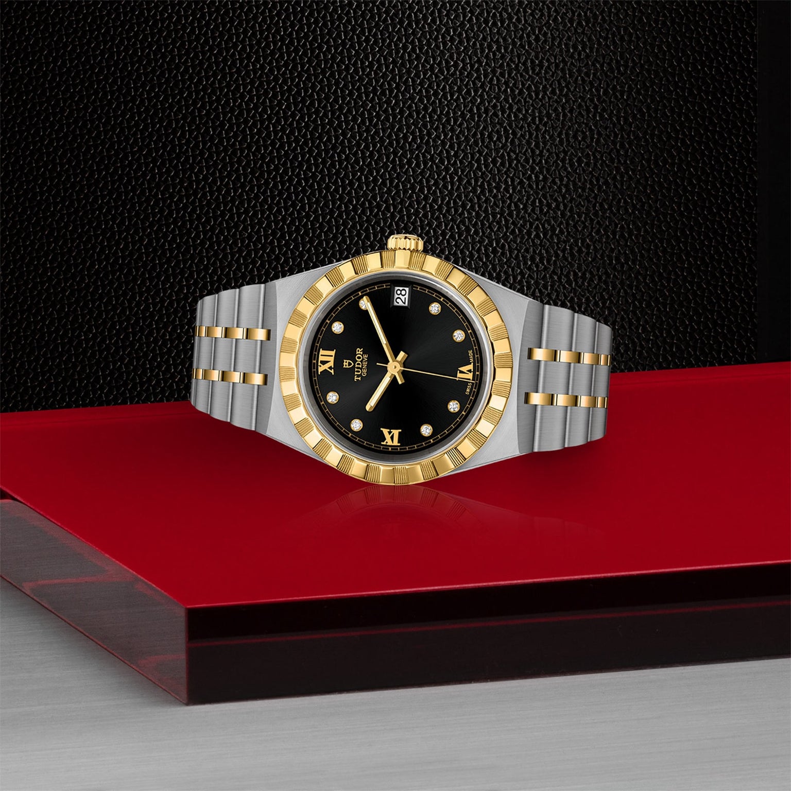 A TUDOR Royal wristwatch in two-tone gold and silver with a black dial, Roman numerals, and diamond hour markers rests on a red and gray surface, reflecting the brand’s automatic sport-chic style and technical excellence.