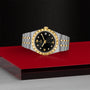 A TUDOR Royal wristwatch in two-tone gold and silver with a black dial, Roman numerals, and diamond hour markers rests on a red and gray surface, reflecting the brand’s automatic sport-chic style and technical excellence.
