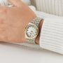 A wrist wearing the TUDOR Royal watch in stainless steel and gold with a white dial, Roman numerals, and date display, paired with a white knitted sweater. Ideal for those who love automatic sport-chic timepieces.