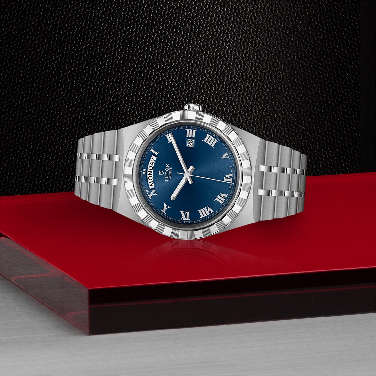 The TUDOR Royal wristwatch, crafted in stainless steel with a blue dial and Roman numerals, features a day-date display and integrated bracelet. Set on red and gray surfaces with a dark background, it radiates sport-chic elegance.