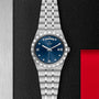 A TUDOR Royal wristwatch in silver with a blue dial, diamond markers, day-date feature, and integrated bracelet, showcased on a textured black and metallic background.