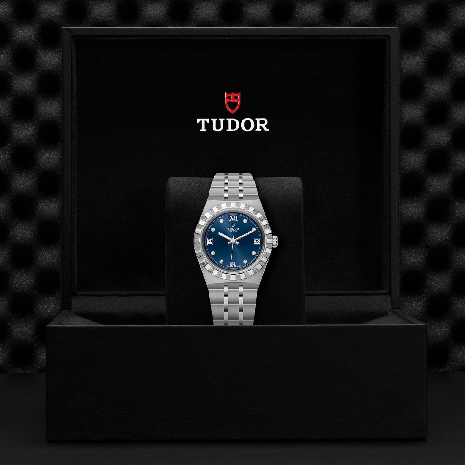 A TUDOR Royal wristwatch with a blue dial is showcased on a stand inside an open black Tudor watch box, highlighting the refined elegance of this automatic sport-chic timepiece.