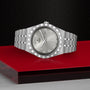 A TUDOR Royal wristwatch in silver, featuring an integrated bracelet, Roman numeral markers, and day-date displays, is displayed on a red and black geometric surface.