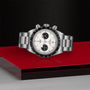 The TUDOR Black Bay Chrono, a stainless steel wristwatch with a white dial, black subdials, and tachymeter bezel, is displayed on a red and gray geometric surface—highlighting the brand’s signature chronograph style.