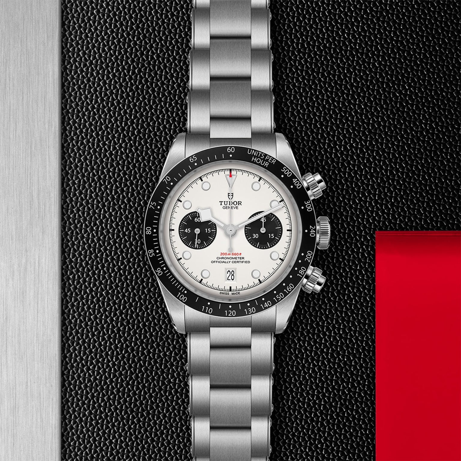 A TUDOR Black Bay Chrono with a stainless steel case, white dial, black subdials, and metal bracelet is showcased on a textured black and red surface.